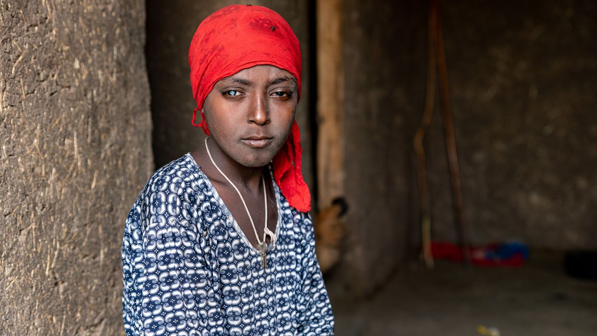 Auf dem Bild ist Mulunesh abgebildet. Sie trägt ein rotes Tuch um den Kopf und ein blaues Kleid. Sie sitzt in einer Hütte.