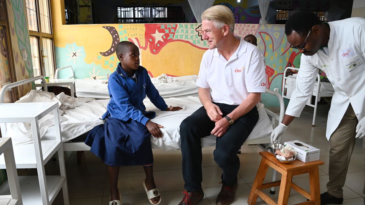 Ein Mann in weißem Polo-Hemd und Jeans und ein Mädchen sitzen auf der Kante eines Krankenbettes in einem Krankenzimmer. Ein weiterer Mann stellt Medizinprodukte auf einen Stuhl neben dem Bett.