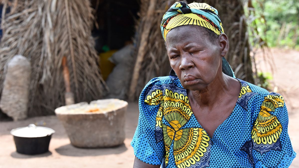 Eine etwa sechzigjährige Frau im blau-gelben Baumwollkleid und buntem Kopftuch sitzt mit geschlossenen Augen vor einer einfachen, mit trockenen Palmwedeln bedeckten Hütte.