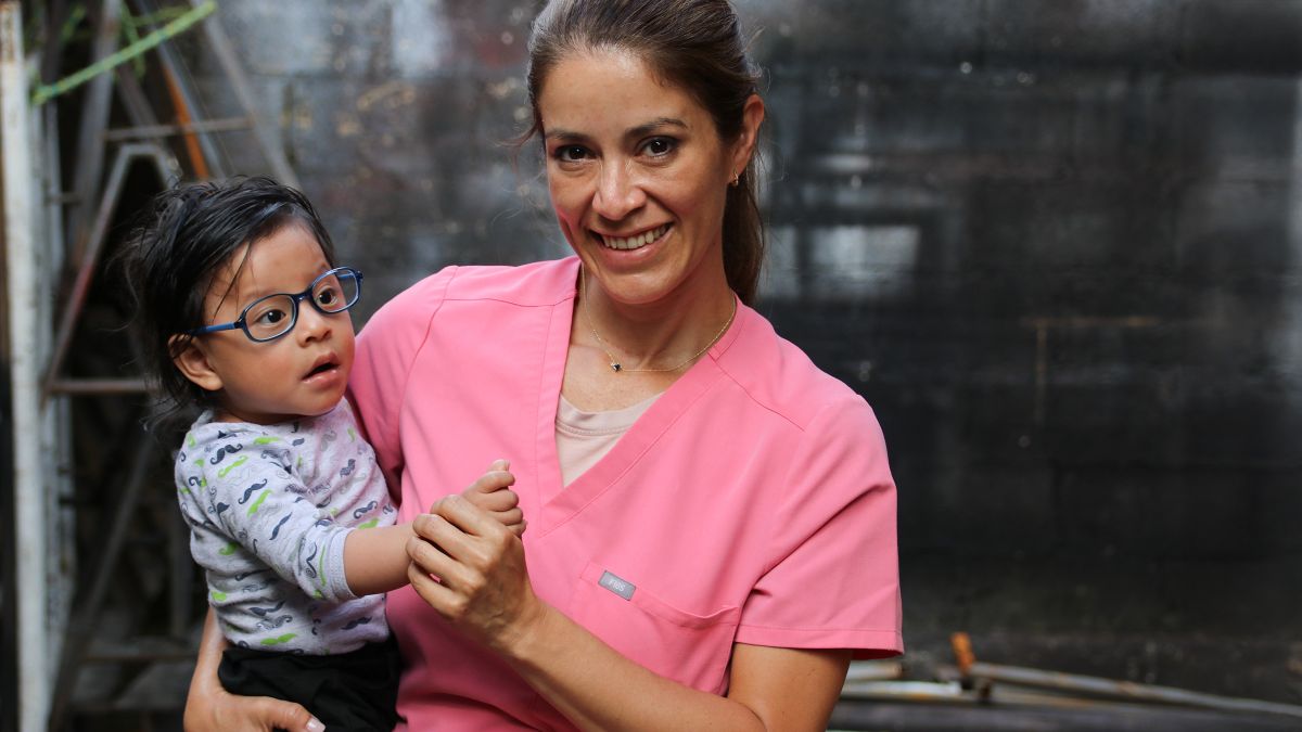 Eine Frau in rosa Kittel hält ein Kleinkind mit Brille auf dem Arm.