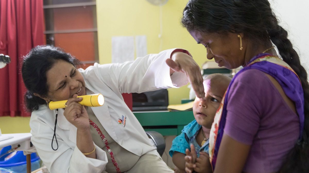 Eine Frau im Arztkittel leuchtet mit einer Lampe in die Augen eines Kleinkindes. Es sitzt auf dem Schoß einer Frau mit Sari.