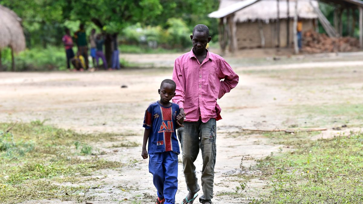 Ein etwa neunjähriger Junge führt einen Mann Mitte vierzig an der Hand. Im Hintergrund sind Hütten zu sehen.