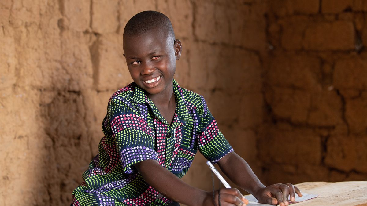 Jovithe sitzt an einen Tisch mit einem Stift in der Hand und lächelt den Betrachter an.