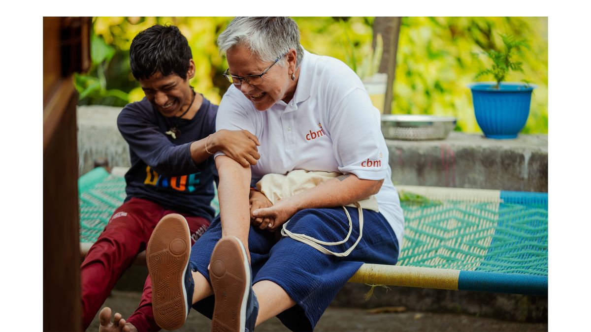 Eine Frau mit grauem Kurzhaarschnitt und ein Teenagerjunge sitzen lachend nebeneinander auf einer Liege mit bunter, gewebter Bespannung.
