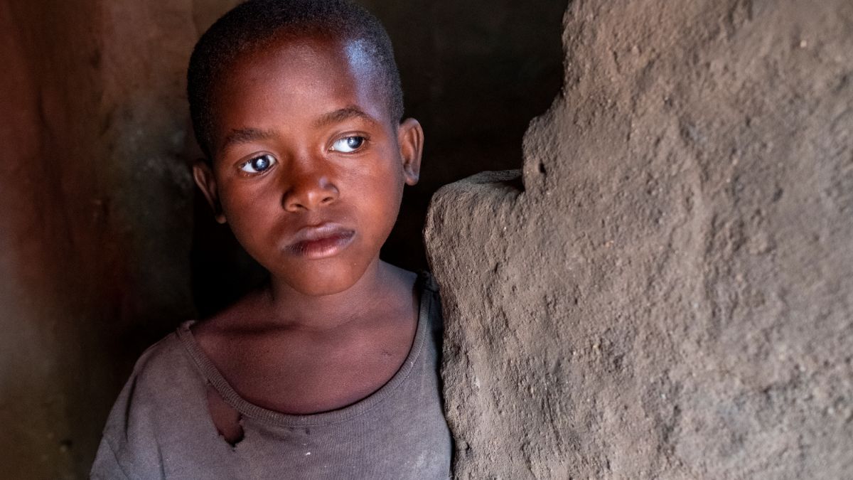Ein Mädchen mit sehr kurzem Haar und weißen Pupillen steht an einer Lehmwand in einem dunklen Hüttenraum.