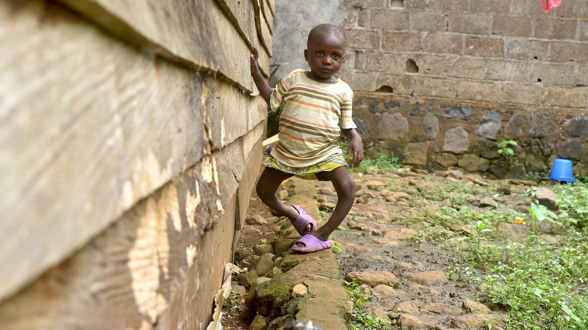 Ein etwa fünfjähriges Mädchen geht an einer Holzhütte auf steinigem Boden entlang.