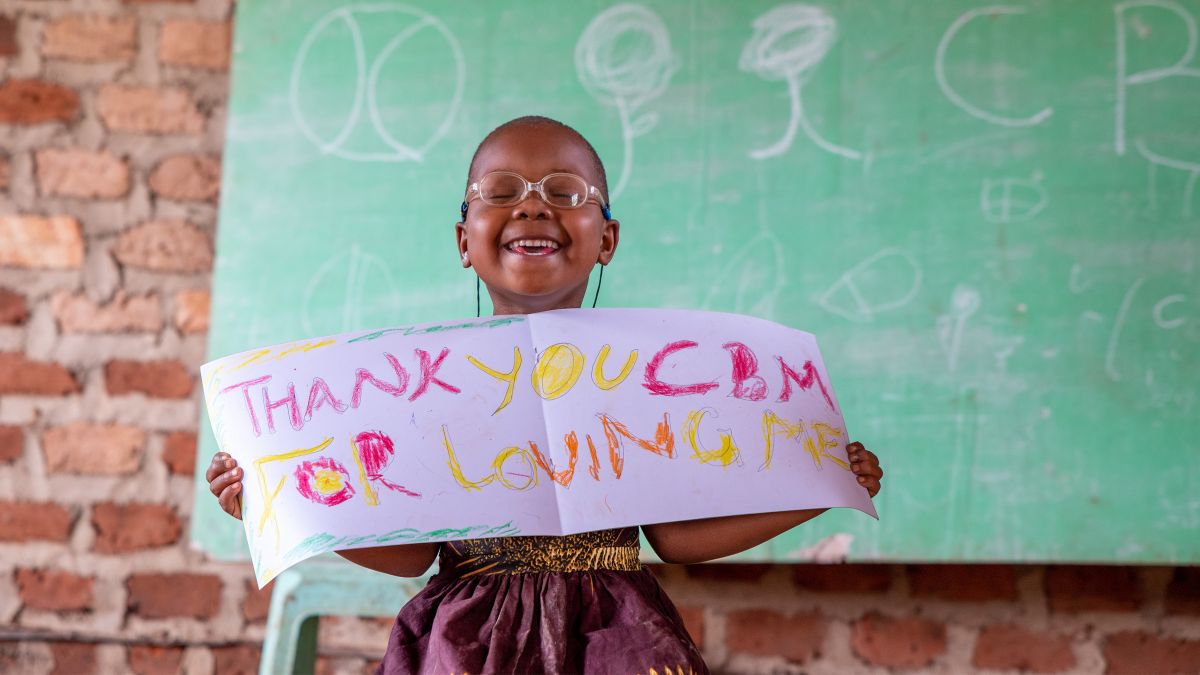 Ein lachendes kleines Mädchen mit Brille hält ein Blatt, auf dem steht: Thank you cbm.