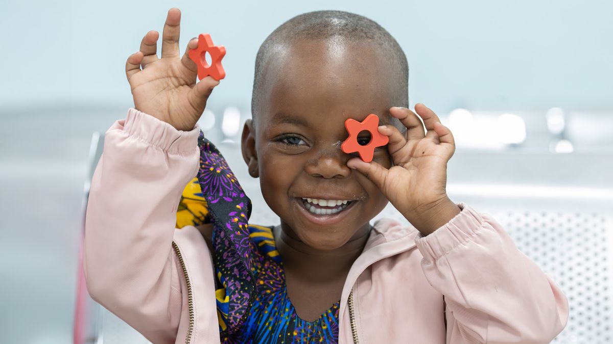 Ein kleines Mädchen lacht und hält eine rote Holz- oder Plastik-Blume vor sein linkes Auge. In der rechten Hand hält es eine zweite Blume hoch.