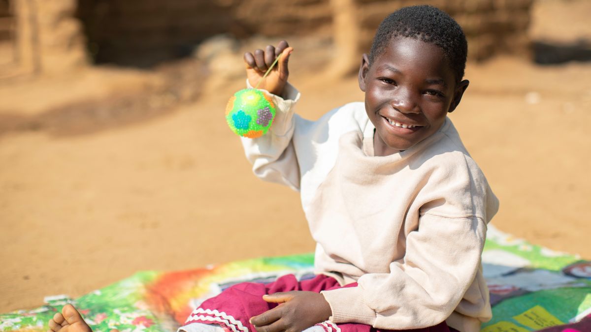 Lächelndes Mädchen aus Malawi sitzt auf dem Boden und hält einen bunten Ball hoch
