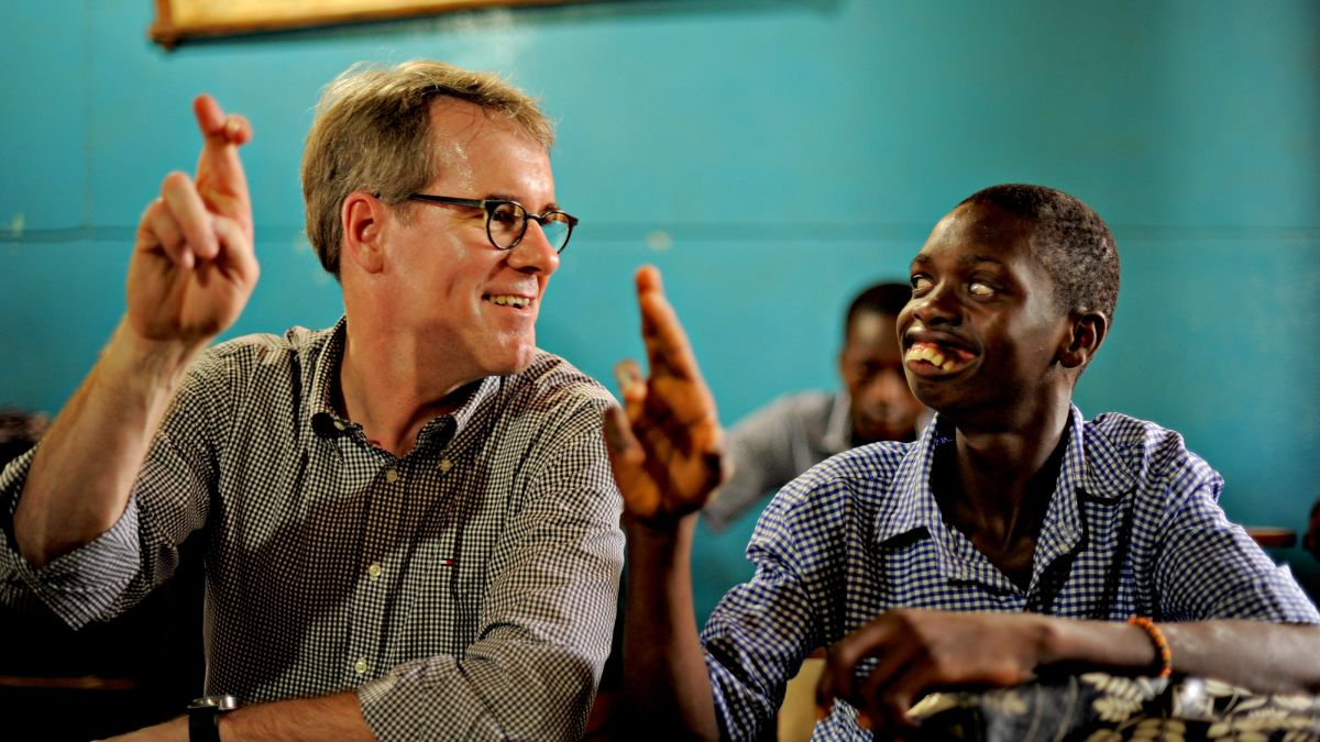 Ein Mann mit Brille und ein afrikanischer Junge sitzen an einem Schulpult und machen das gleiche Fingerzeichen.
