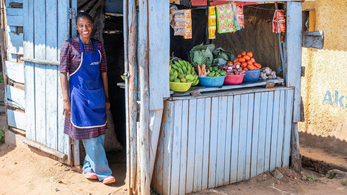 Eine junge Frau mit Schürze steht neben einer kleinen, blaugestrichenen Bretterbude, auf deren Auslage Obst und Gemüse liegen.