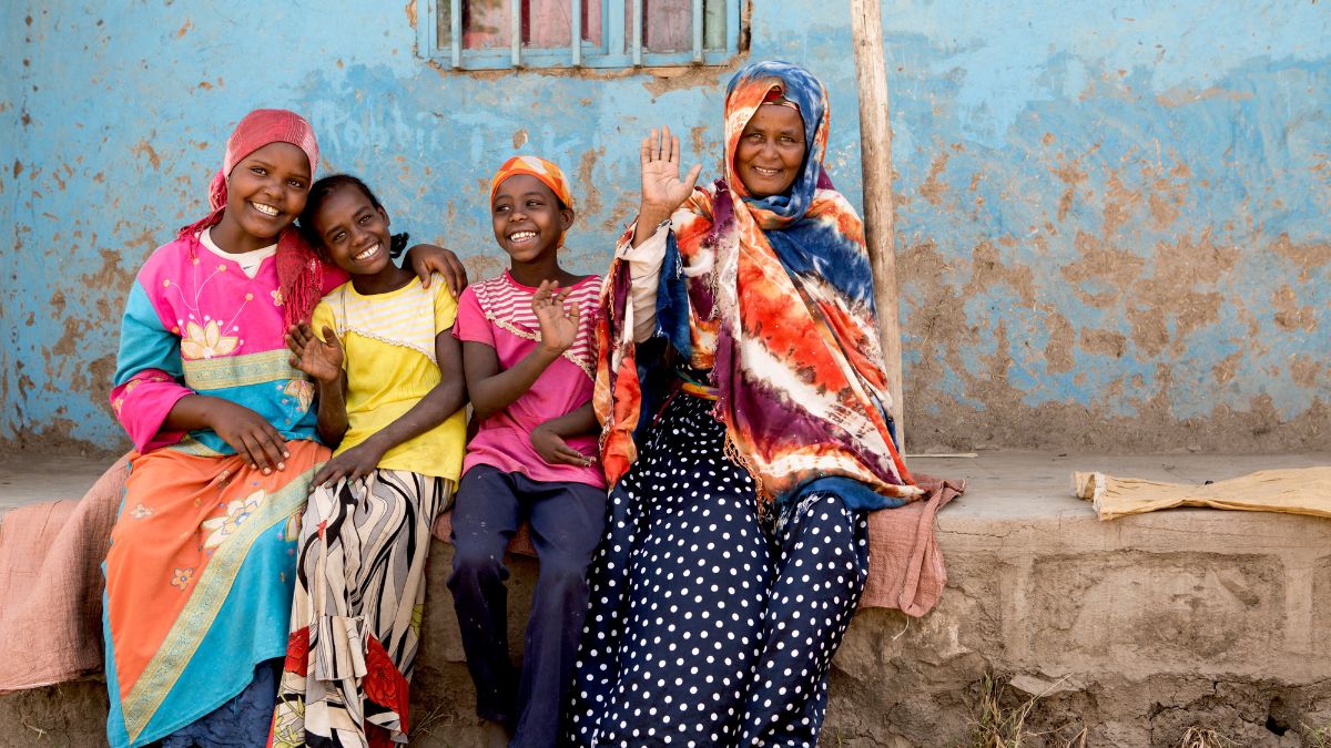 Zwei Frauen und zwei Mädchen sitzen lächelnd auf einem Mauervorsprung und winken.