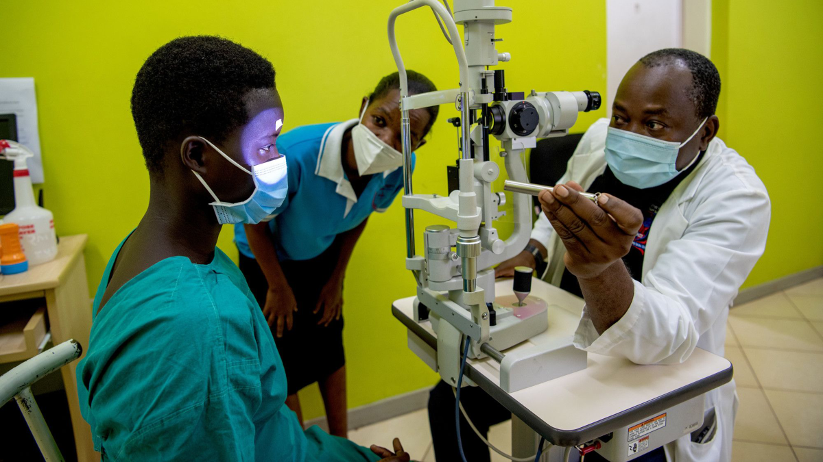 Ein afrikanischer Mann im weißen Kittel mit Mund-Nasen-Schutz leuchtet ins Auge eines Mädchens, um dessen Auge zu untersuchen.