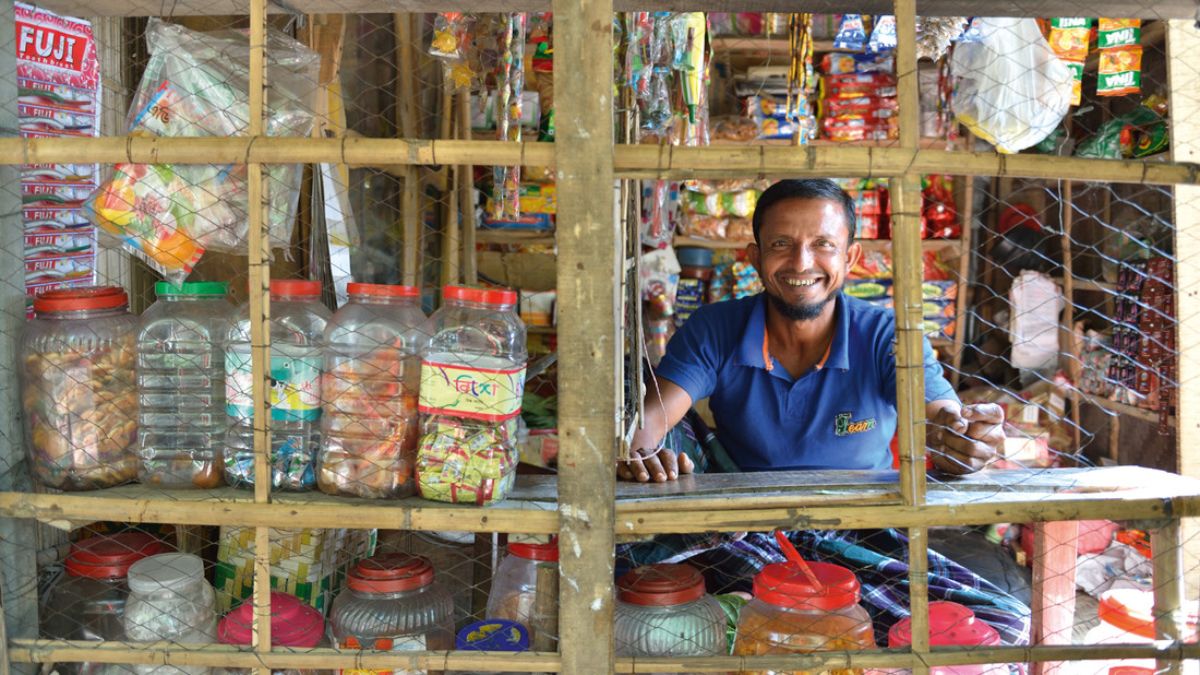 Ein Mann sitzt lachend an der Theke eines Kiosks. Neben ihm stehen große Gläser mit Lebensmitteln und Süßigkeiten.
