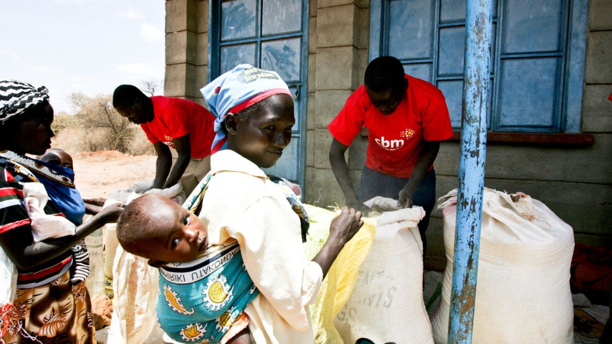 Zwei afrikanische Frauen mit Babys auf dem Rücken stehen mit leeren Tüten vor zwei afrikanischen Männern, die vor zwei vollen Säcken stehen.