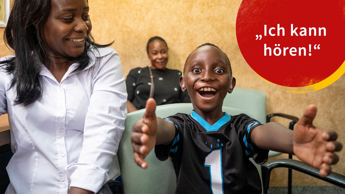 Ein Junge mit Hörgeräten streckt die Arme aus, eine Frau in weißer Bluse sitzt neben ihm, eine weitere Frau hinter ihm an der Wand. Alle lachen.