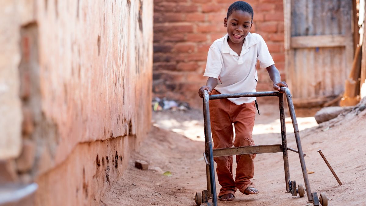 Ein afrikanischer Junge läuft mit einer Gehhilfe an einer Hauswand entlang.
