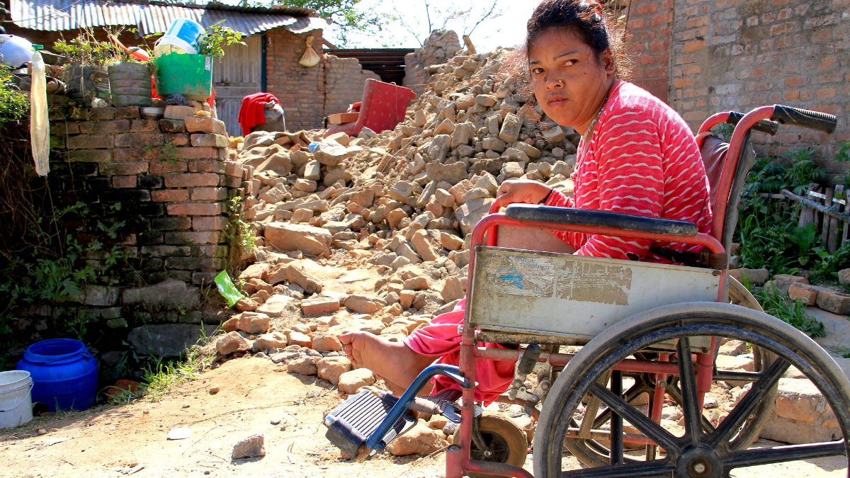 Eine Frau im Rollstuhl sitzt neben einem zerstörten Haus.