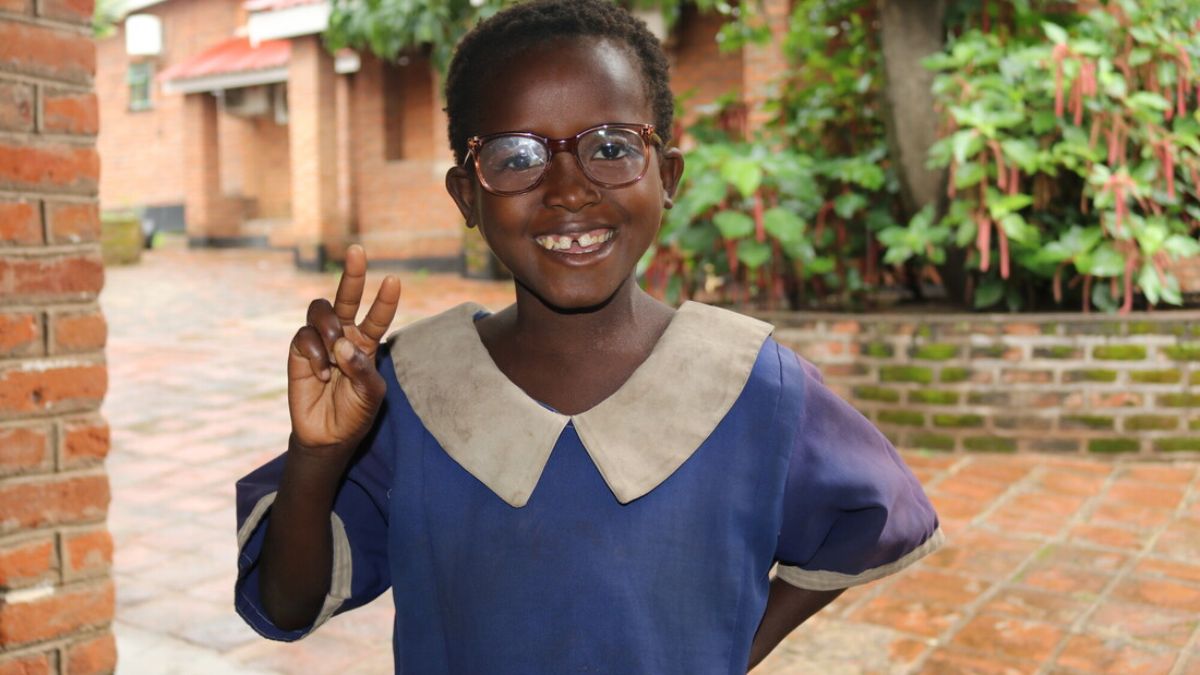 Ein Mädchen mit Brille lächelt und zeigt das Victoryzeichen mit der rechten Hand.