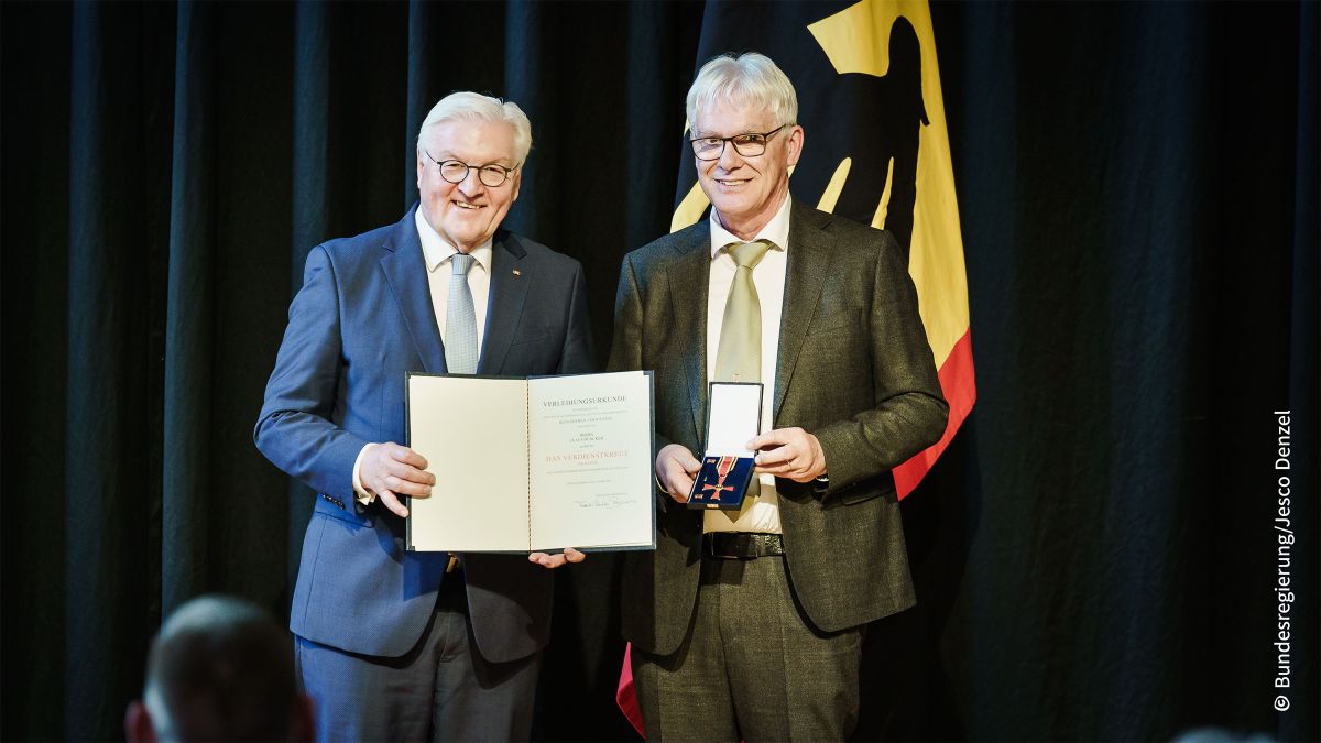 Zwei Männer stehen nebeneinander. Der eine hält eine Urkunde in den Händen, der andere ein Etui mit einer Medaille.