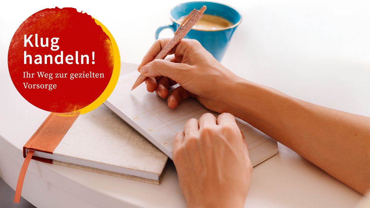 Eine Hand hält einen Stift und schreibt auf einen Block, der auf einer Kladde liegt, daneben steht eine Kaffeetasse.