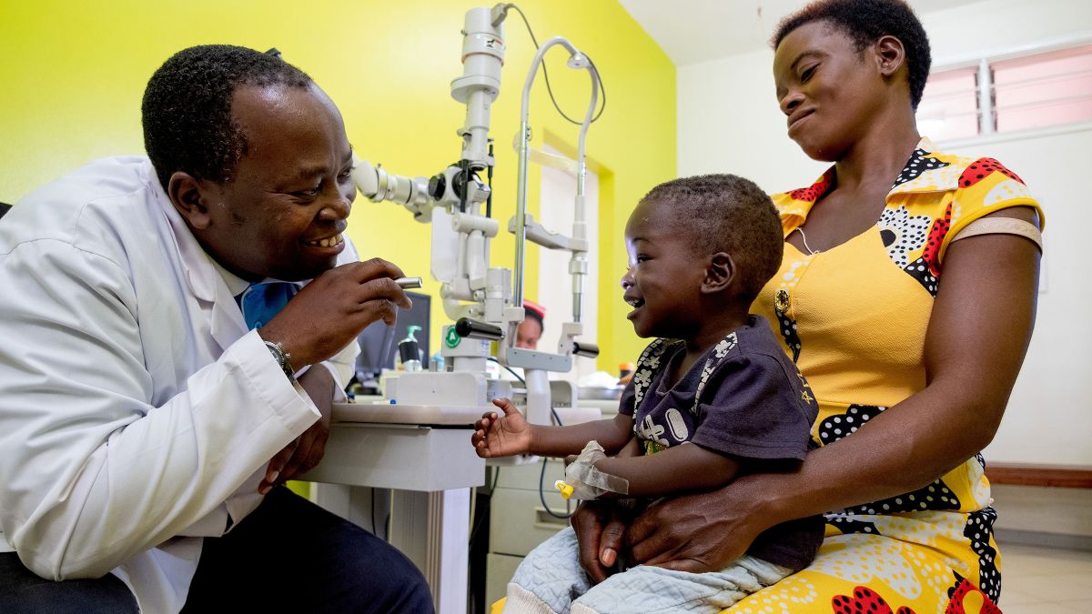 Ein afrikanischer Mann im weißen Kittel leuchtet einem Kleinkind in einer Klinik mit einer Taschenlampe ins Auge. Die Klinik ist Partner der seriösen Spendenorganisation CBM.