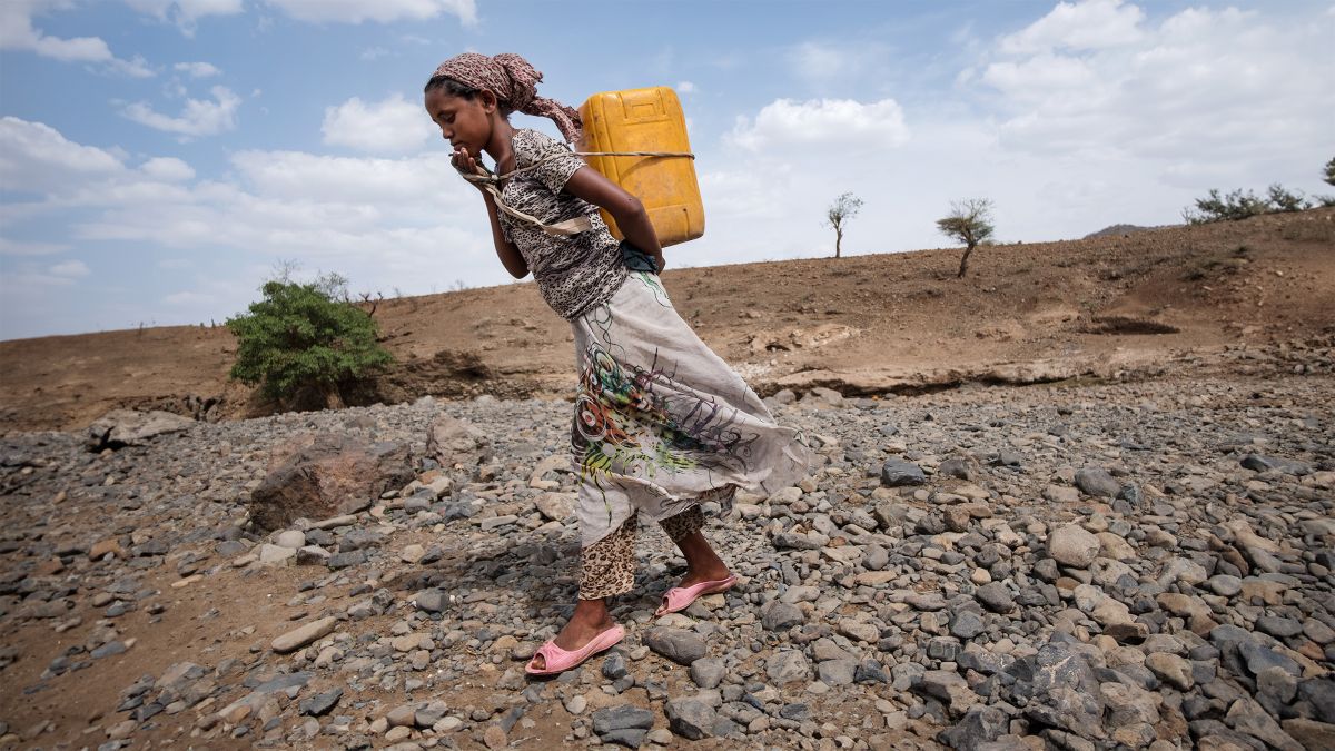 Ein junges Mädchen mit Kopftuch, bunter Bluse und Rock trägt einen Wasserkanister auf dem Rücken. Es geht in Plastikballerinas über einen kiesigen Grund durch ein fast baum- und strauchloses Gelaende.