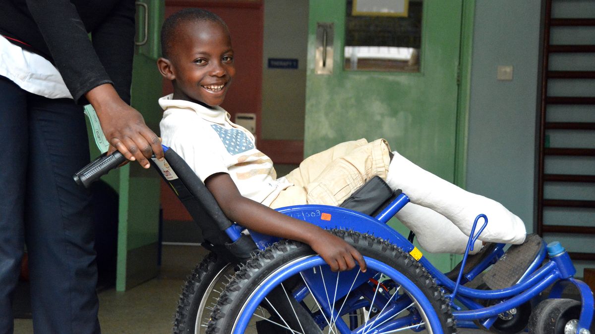 Ein lachender Junge mit Gipsbeinen sitzt im Rollstuhl.