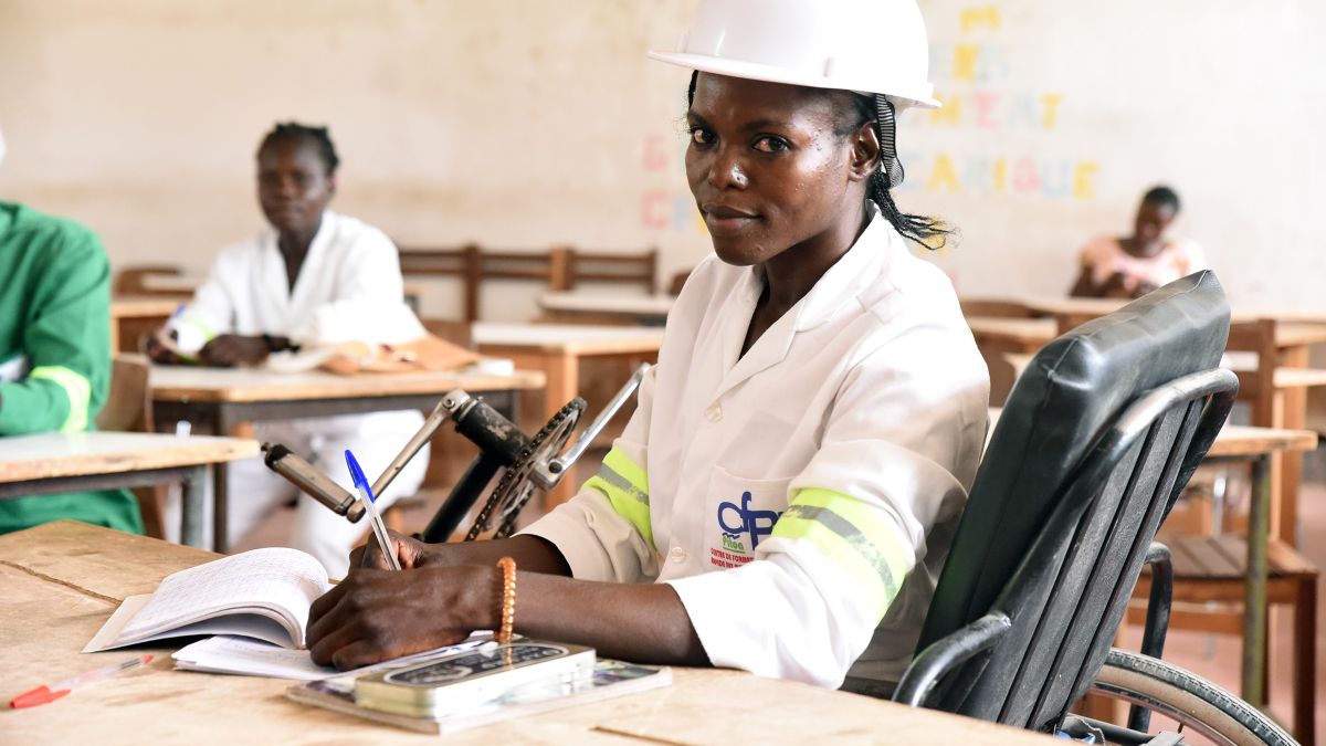 Eine junge Frau im Rollstuhl sitzt am Tisch in einem Klassenraum. Sie trägt einen Schutzhelm.