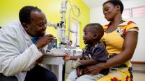 Ein afrikanischer Mann im weißen Kittel leuchtet einem Kleinkind in einer Klinik mit einer Taschenlampe ins Auge. Die Klinik ist Partner der seriösen Spendenorganisation CBM.