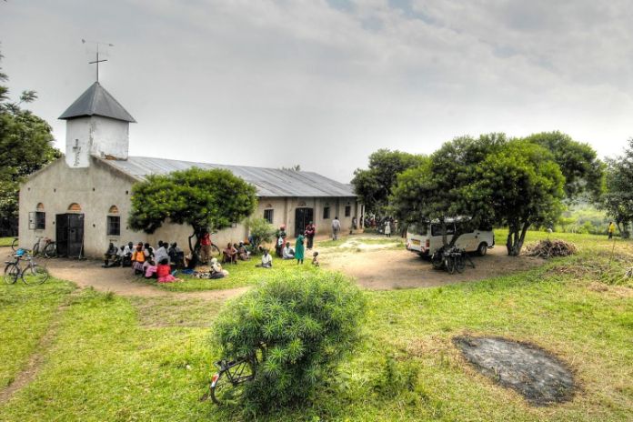 Landschaft mit Kirche, vor der eine Gruppe Menschen in bunter Kleidung sitzt.