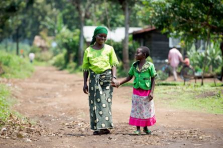 Eine Frau und ein Mädchen laufen Hand in Hand über einen Sandweg im Grünen.