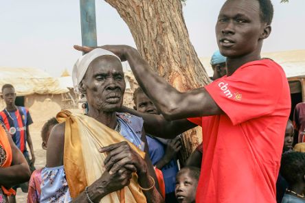 Ein junger Mann im rotem T-Shirt mit CBM-Logo misst mit einer Holzlatte die Körpergröße einer älteren Frau.