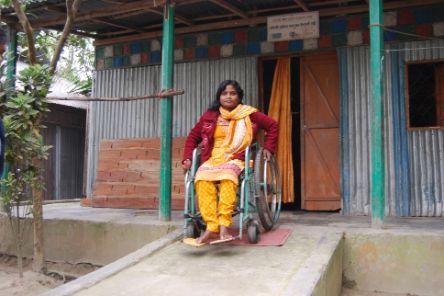 Eine junge Frau aus Bangladesch sitzt im Rollstuhl. Dieser steht auf einer vor einem Haus angebrachten Rampe steht.