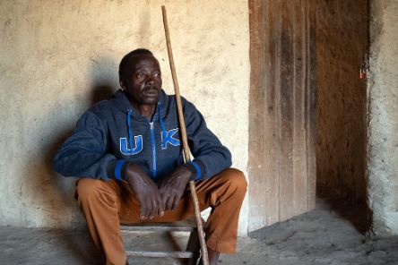 Ein Mann sitzt in einer lehmverputzten Hütte auf einem Holzschemel. Er hält einen Stock in der Hand.