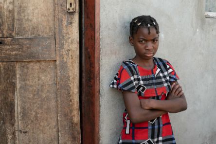 Ein etwa zehnjähiges Mädchen steht mit verschränkten Armen vor einem einfachen kleinen Haus.