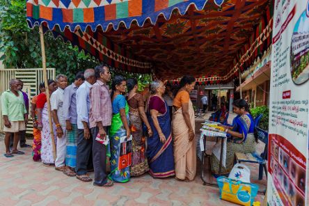 Frauen in Saris und Männer stehen unter einem bunten Stoffbaldachin Schlange.