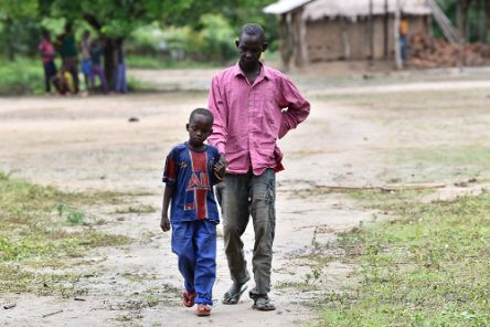 Ein etwa neunjähriger Junge führt einen Mann Mitte vierzig an der Hand. Im Hintergrund sind Hütten zu sehen.