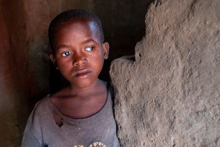 Ein Mädchen mit sehr kurzem Haar und weißen Pupillen steht an einer Lehmwand in einem dunklen Hüttenraum.