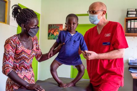 Ein kleines Mädchen mit extremen O-Beinen steht auf einem Untersuchungstisch, links steht eine Frau, rechts ein Mann, die das Kind halten.