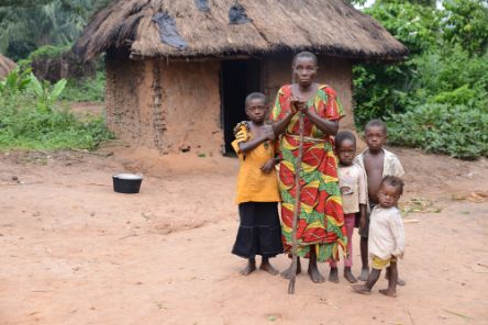 Eine alte blinde Frau mit Stock steht mit mehreren Kindern vor einer Hütte.