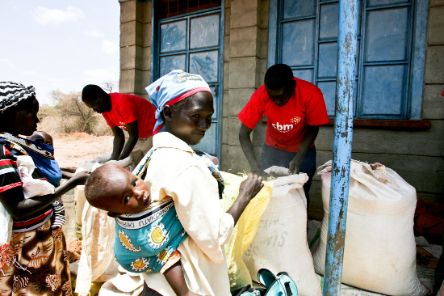 Zwei afrikanische Frauen mit Babys auf dem Rücken stehen mit leeren Tüten vor zwei afrikanischen Männern, die vor zwei vollen Säcken stehen.