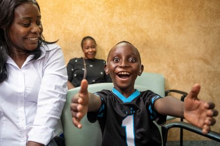 Ein Junge mit Hörgeräten streckt die Arme aus, eine Frau in weißer Bluse sitzt neben ihm, eine weitere Frau hinter ihm an der Wand. Alle lachen.