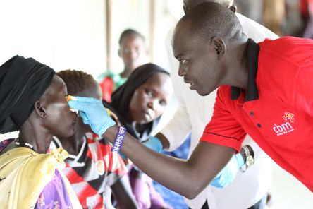 Ein Mann entfernt ein Pflaster vom Auge einer Frau. Andere Personen schauen dabei zu.