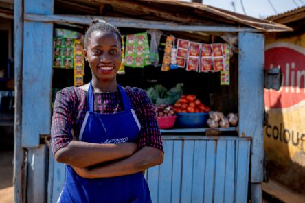 Eine junge Frau steht mit verschränkten Armen vor ihrem Marktstand und lächelt. Das Bild vermittelt, dass sie anpacken kann.