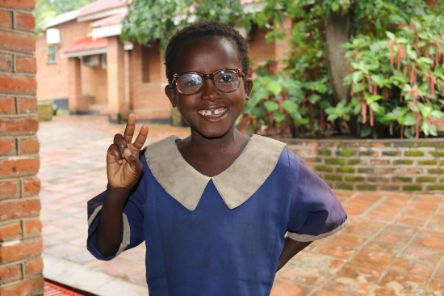 Ein Mädchen mit Brille lächelt und zeigt das Victoryzeichen mit der rechten Hand.