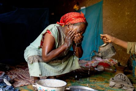 Eine Frau sitzt in der Hocke auf dem Boden und wäscht sich das Gesicht mit Wasser aus einer kleinen Kanne.