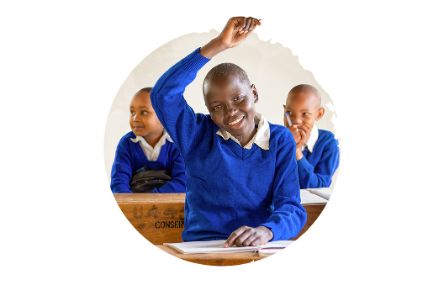 Junge in blau-weißer Schuluniform sitzt in seiner Schulbank im Klassenzimmer und streckt lachend einen Arm hoch, um sich zu melden.