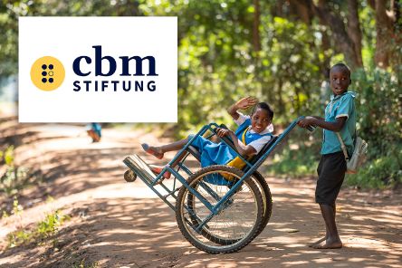 Im Rollstuhl sitzendes Mädchen lacht und winkt. Ein Junge schiebt den Rollstuhl.