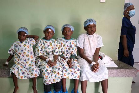 Vier Kinder im Alter von etwa fünf bis 14 Jahren sitzen nebeneinander. Sie tragen OP-Kleidung und haben jeweils ein Pflaster auf der Stirn.